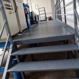 MS STEEL CONSTRUCTION SPÓŁKA Z OGRANICZONĄ ODPOWIEDZIALNOŚCIĄ - Metalowa platforma robocza z kratownicowymi schodami w hali produkcyjnej, widoczna szafa sterownicza i urządzenia przemysłowe w tle.