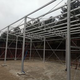 MS STEEL CONSTRUCTION SPÓŁKA Z OGRANICZONĄ ODPOWIEDZIALNOŚCIĄ - Konstrukcja stalowa z profili zamkniętych, przygotowana do dalszej zabudowy, widoczne rusztowania i drzewa w tle.