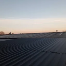 Nowa, ciemnoszara blacha trapezowa na dachu hali przemysłowej, widoczna konstrukcja stalowa, w tle starsze pokrycie dachowe i kominy wentylacyjne, niebo o zmierzchu.