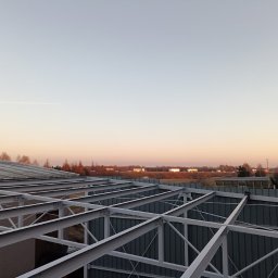 MS STEEL CONSTRUCTION SPÓŁKA Z OGRANICZONĄ ODPOWIEDZIALNOŚCIĄ - Konstrukcja stalowa budynku w trakcie montażu, widok z góry na szkielet z profili pomalowanych na szaro, w tle zachodzące słońce i krajobraz podmiejski z widocznymi samochodami na drodze.