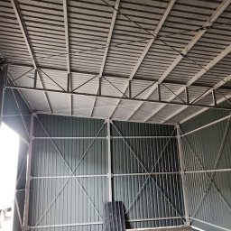 MS STEEL CONSTRUCTION SPÓŁKA Z OGRANICZONĄ ODPOWIEDZIALNOŚCIĄ - Wnętrze szkieletowej konstrukcji hali z blachy trapezowej, widoczne wzmocnienia krzyżowe i otwór wejściowy z widocznym fragmentem pojazdu.