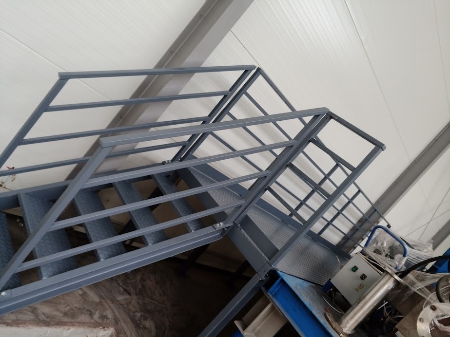 Schody przemysłowe z szarą metalową konstrukcją i zabezpieczeniami w hali produkcyjnej, widoczne elementy maszyny w tle.