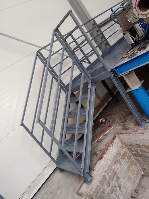 Metalowe schody z platformą roboczą w szarej kolorystyce, prowadzące do urządzenia przemysłowego, widoczne na tle białej ściany i betonowej posadzki z fragmentami cegieł