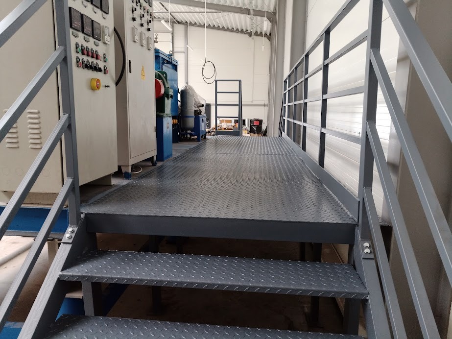 Metalowa platforma robocza z schodami i barierkami, powierzchnia z blachy ryflowanej, w tle maszyny przemysłowe i regał.