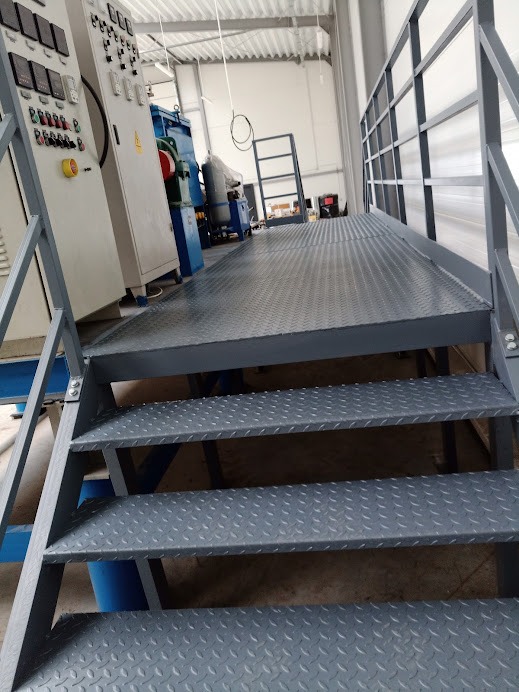 Metalowa platforma robocza z kratownicowymi schodami w hali produkcyjnej, widoczne maszyny przemysłowe w tle.