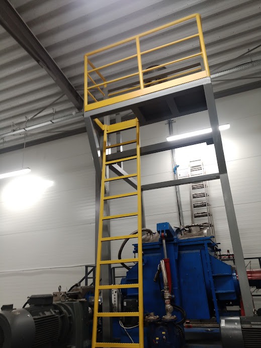 Żółta drabina i platforma z barierami ochronnymi przy maszynie przemysłowej w hali produkcyjnej.