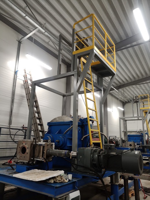 Wnętrze hali przemysłowej z niebieskim mieszalnikiem przemysłowym na stalowej platformie z żółtymi schodami i balustradami, w tle widoczne podobne urządzenia.