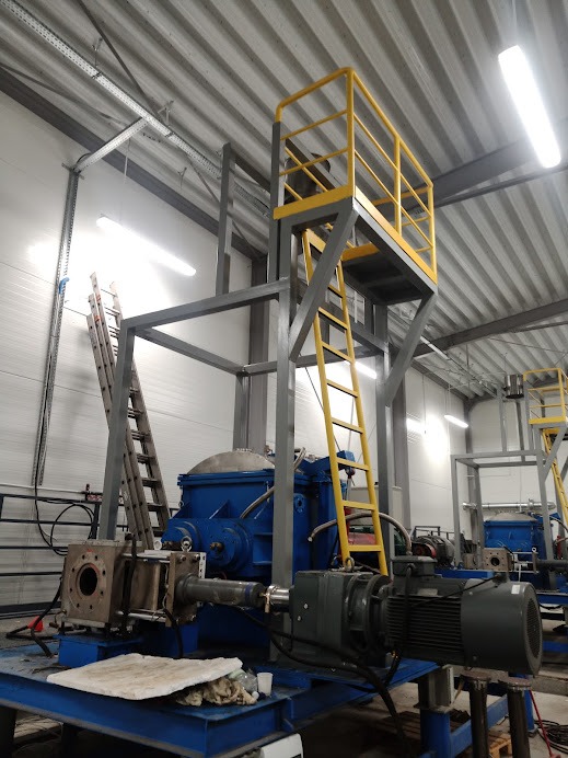 Wnętrze hali produkcyjnej z niebieską maszyną przemysłową na platformie z żółtą drabinką i barierkami ochronnymi, widoczne instalacje i oświetlenie sufitowe.