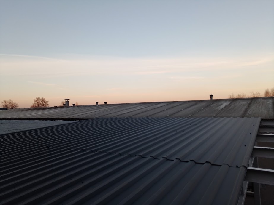 Nowa, ciemnoszara blacha trapezowa na dachu hali przemysłowej, widoczna konstrukcja stalowa, w tle starsze pokrycie dachowe i kominy wentylacyjne, niebo o zmierzchu.