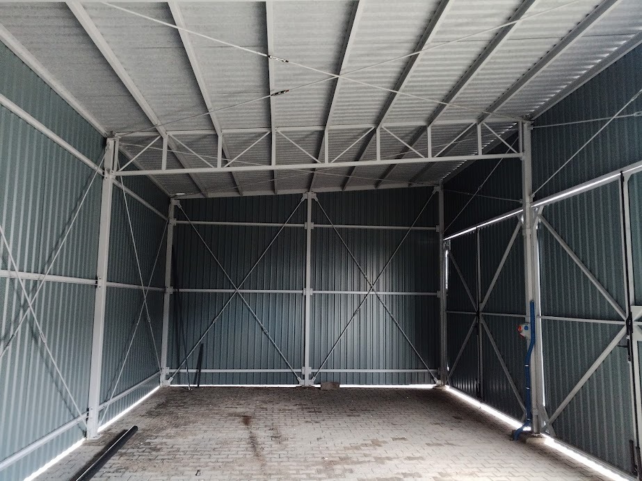 Wnętrze szkieletowej konstrukcji stalowej z blachy falistej, widok na pustą przestrzeń z ceglaną podłogą i metalowym dachem z poprzecznymi wzmocnieniami.