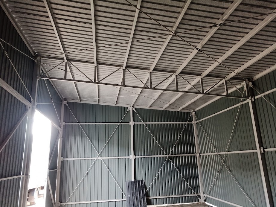 Wnętrze szkieletowej konstrukcji hali z blachy trapezowej, widoczne wzmocnienia krzyżowe i otwór wejściowy z widocznym fragmentem pojazdu.