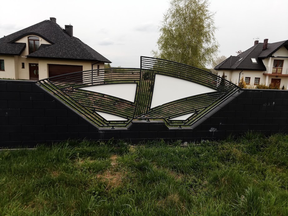 Nowoczesne, czarne ogrodzenie z metalowymi, poziomymi prętami i białymi, trójkątnymi wypełnieniami, na tle domów jednorodzinnych i zielonej trawy.