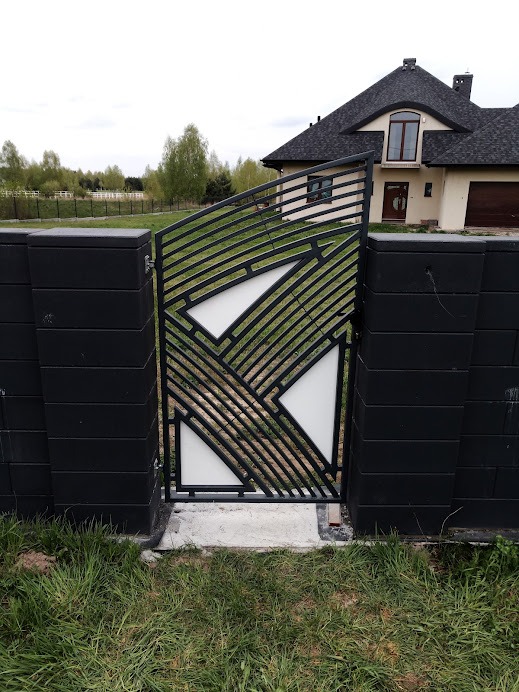 Nowoczesna brama metalowa z ukośnymi prętami i białymi panelami, wkomponowana w czarne, betonowe słupki ogrodzeniowe, na tle domu z ciemnym dachem