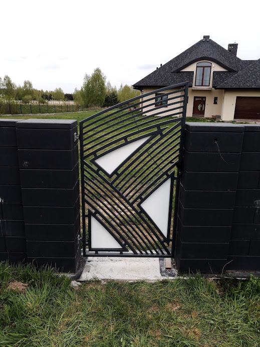 Nowoczesna brama wjazdowa z metalowymi profilami i białymi wypełnieniami, osadzona w czarnych, betonowych słupkach, na tle domu z ciemnym dachem i zielenią.