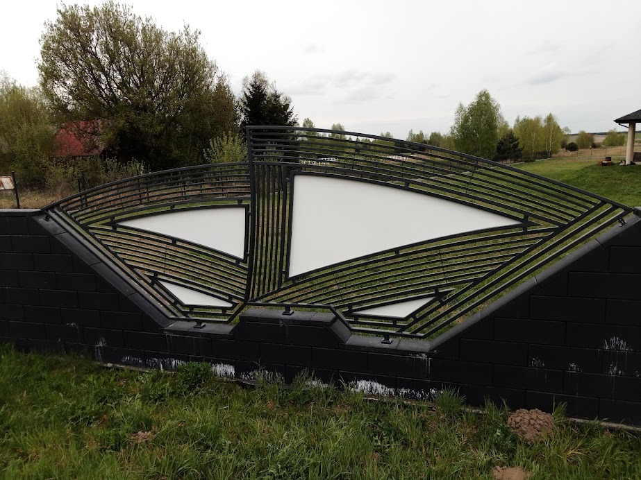 Nowoczesne ogrodzenie z czarnej cegły z metalową konstrukcją o geometrycznym wzorze i białymi wypełnieniami na tle zieleni.