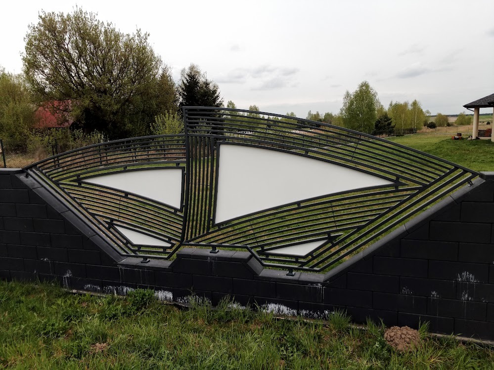 Nowoczesne ogrodzenie z czarnych bloczków betonowych i metalowej konstrukcji z białymi wypełnieniami, w tle drzewa i dom