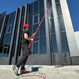 Mężczyzna w czarnej koszulce i czerwonej czapce czyści okna biurowca za pomocą teleskopowej lancy z dyszą, widoczne ślady wody na oknach i czerwony wąż doprowadzający wodę.