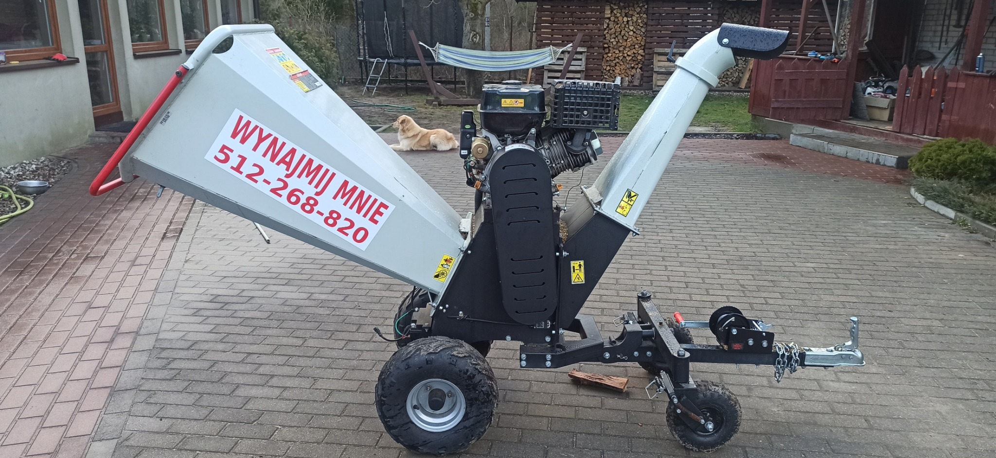 Mobilny rębakiem do gałęzi z napisem 'Wynajmij mnie' na tle kostki brukowej, z psem leżącym w tle i stosem drewna.
