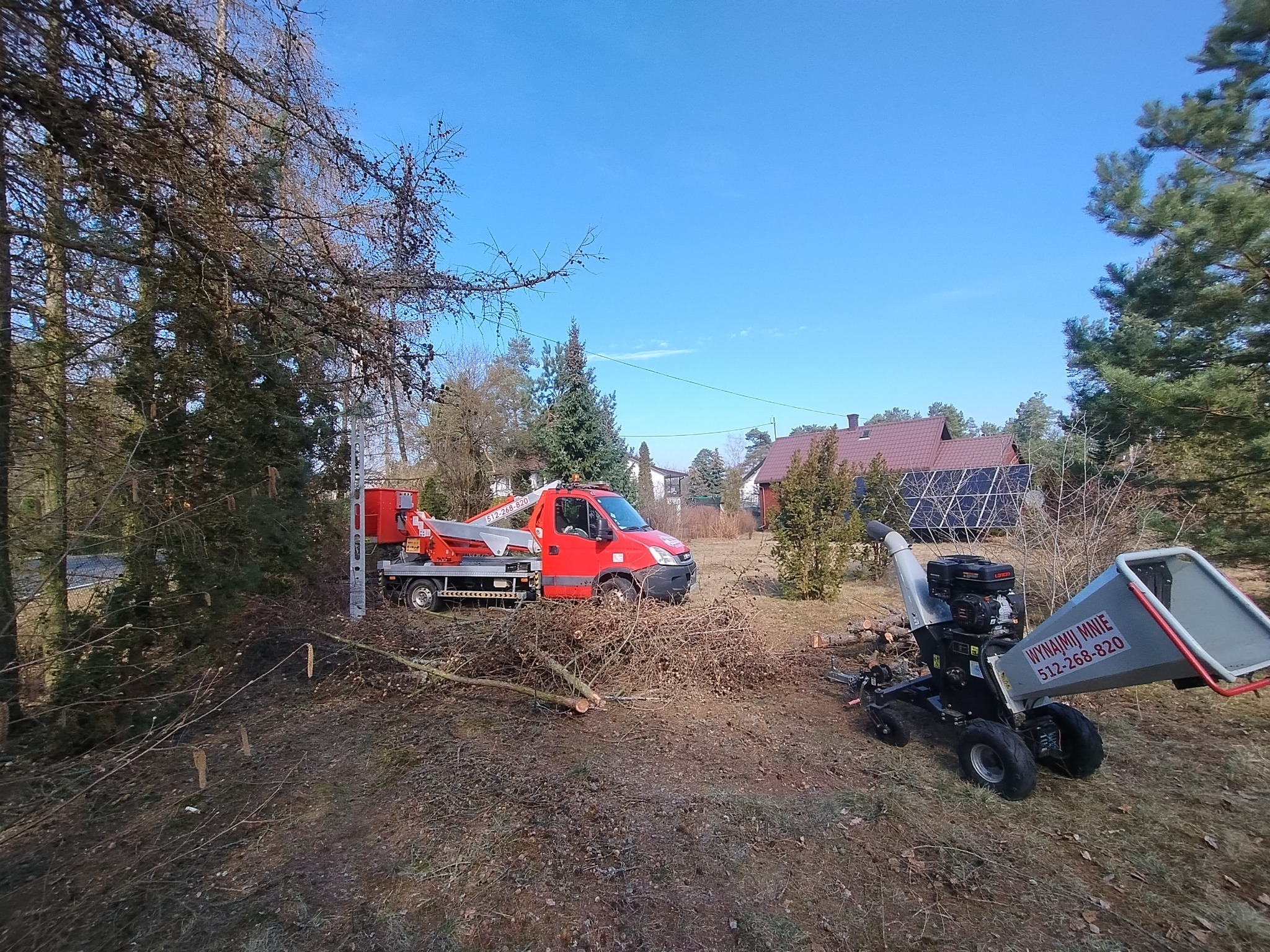 Czerwony podnośnik koszowy i rozdrabniacz gałęzi w Iłach, po wycince. Widoczne ścięte pnie drzew i gałęzie. Błękitne niebo w tle.