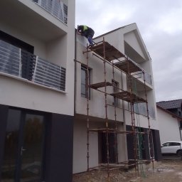 firma budowlana - Pracownik na rusztowaniu wygładza elewację budynku wielorodzinnego z balkonami zabezpieczonymi folią, widoczne ślady prac budowlanych na gruncie.