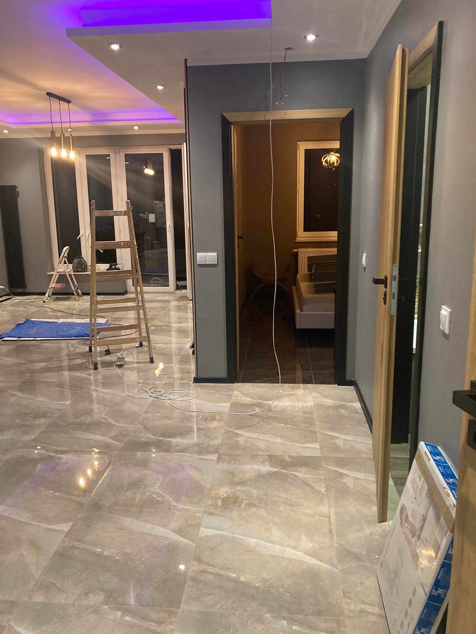 Wnętrze w trakcie prac wykończeniowych: widoczne płytki podłogowe imitujące marmur, drabina, przewody elektryczne zwisające z sufitu, częściowo zamontowane drzwi wewnętrzne z czarną ościeżnicą...