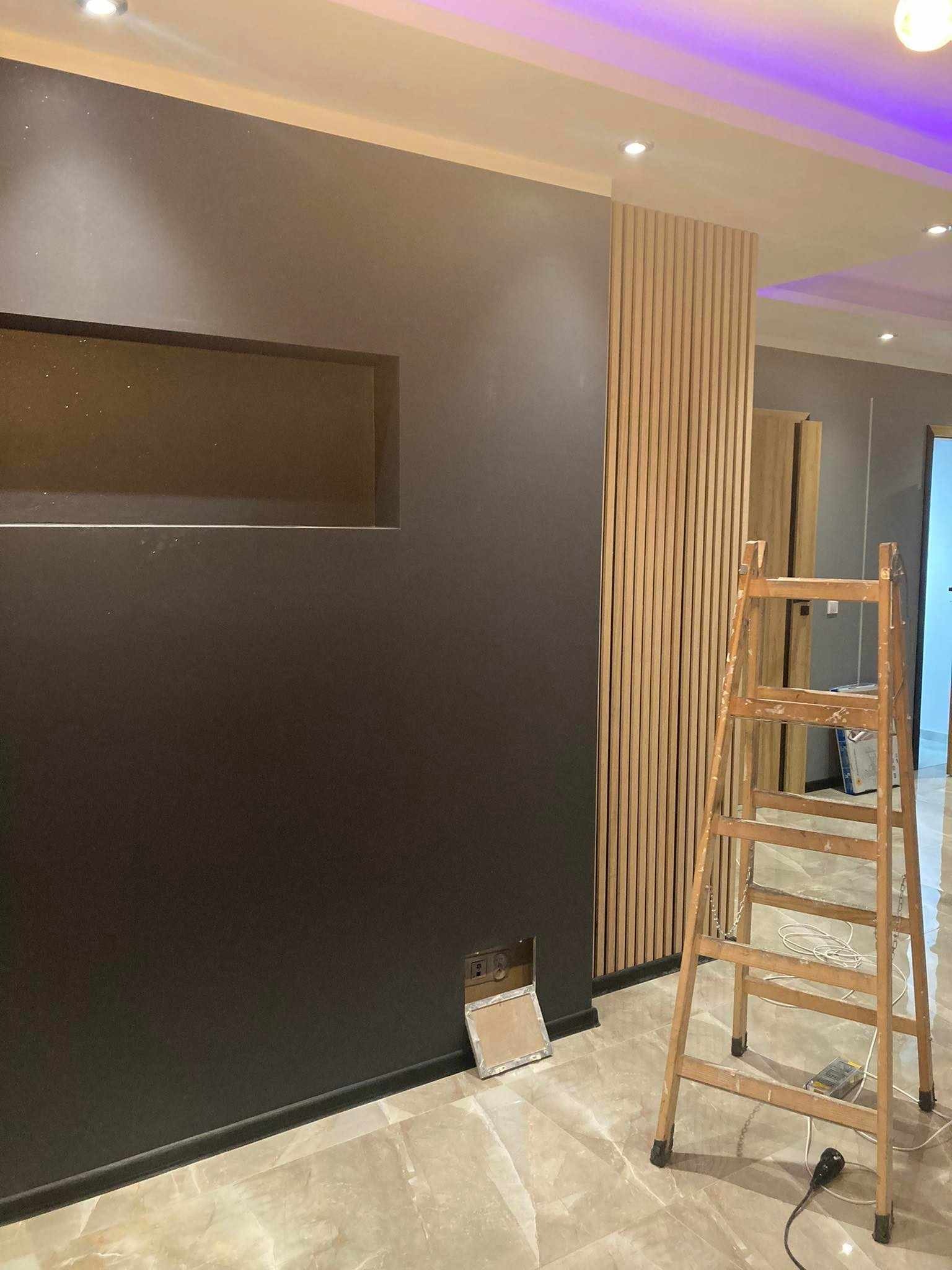 Ciemna ściana z wnęką i drewnianymi panelami dekoracyjnymi, widoczna drabina malarska i przygotowane gniazdko elektryczne, podłoga wyłożona marmurem.