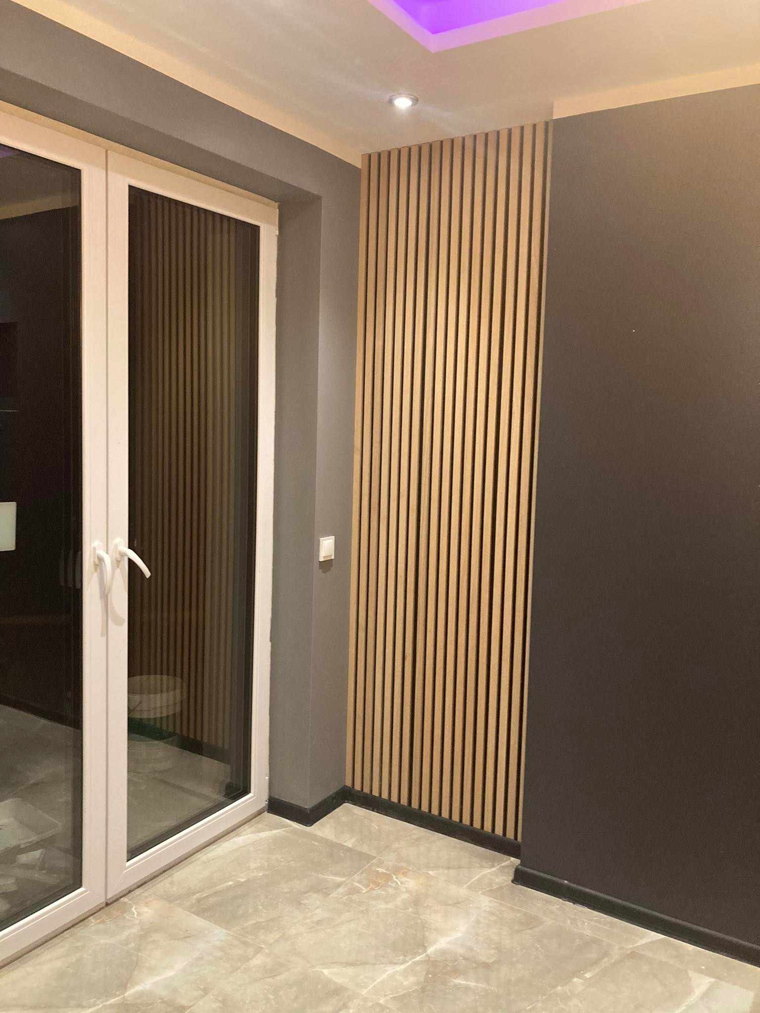 Nowoczesne wnętrze: pionowe drewniane lamele na ścianie w odcieniach beżu, szara ściana obok, marmurowe płytki podłogowe, białe drzwi balkonowe, oświetlenie sufitowe z fioletową poświatą.