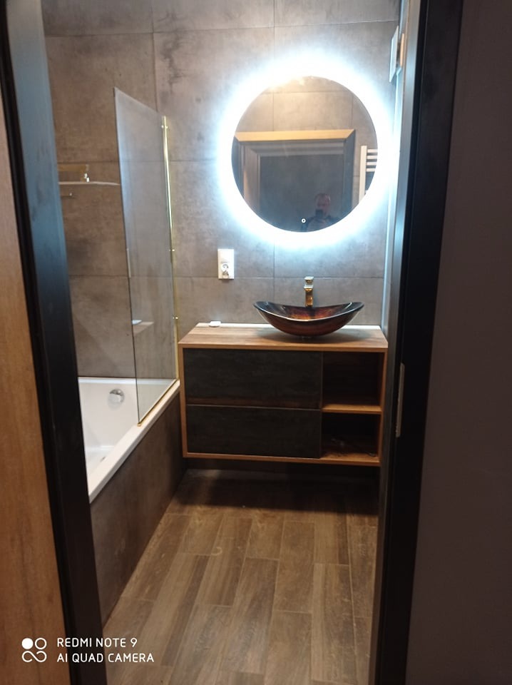 Nowoczesna łazienka z drewnianą podłogą imitującą deski, umywalką w kolorze bursztynu na drewnianej szafce z ciemnymi szufladami, okrągłym lustrem podświetlanym LED i wanną za szklanym panelem.