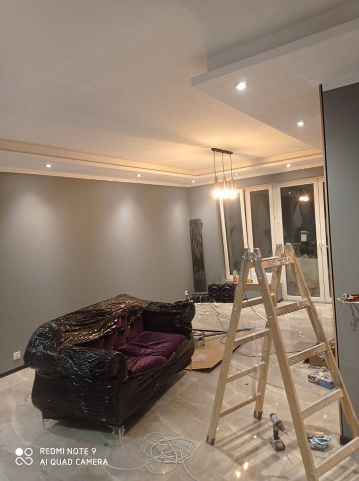 Szare ściany w trakcie remontu mieszkania, zabezpieczona folią kanapa, drewniana drabina i narzędzia na błyszczącej podłodze.