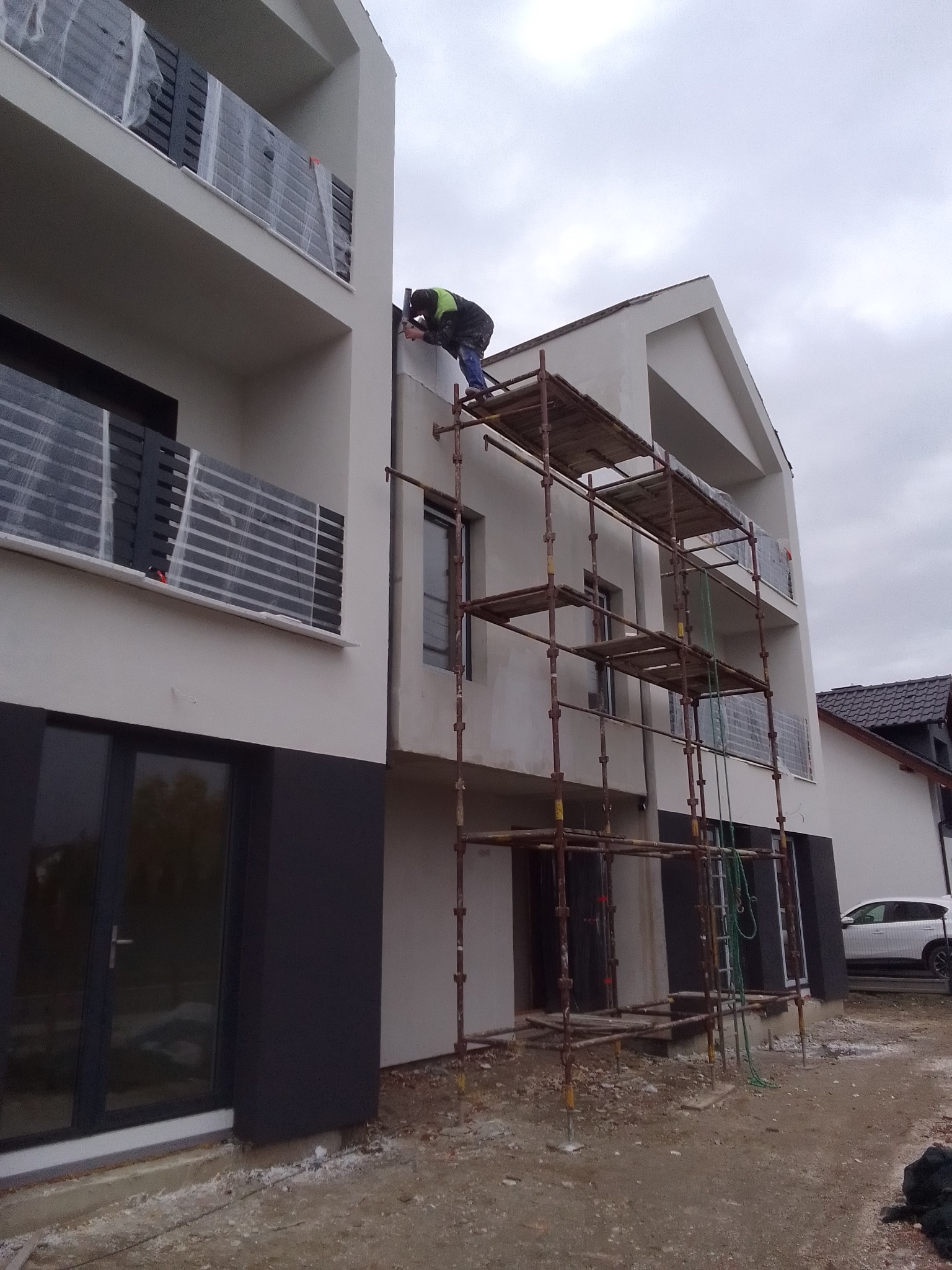 Pracownik na rusztowaniu wygładza elewację budynku wielorodzinnego z balkonami zabezpieczonymi folią, widoczne ślady prac budowlanych na gruncie.