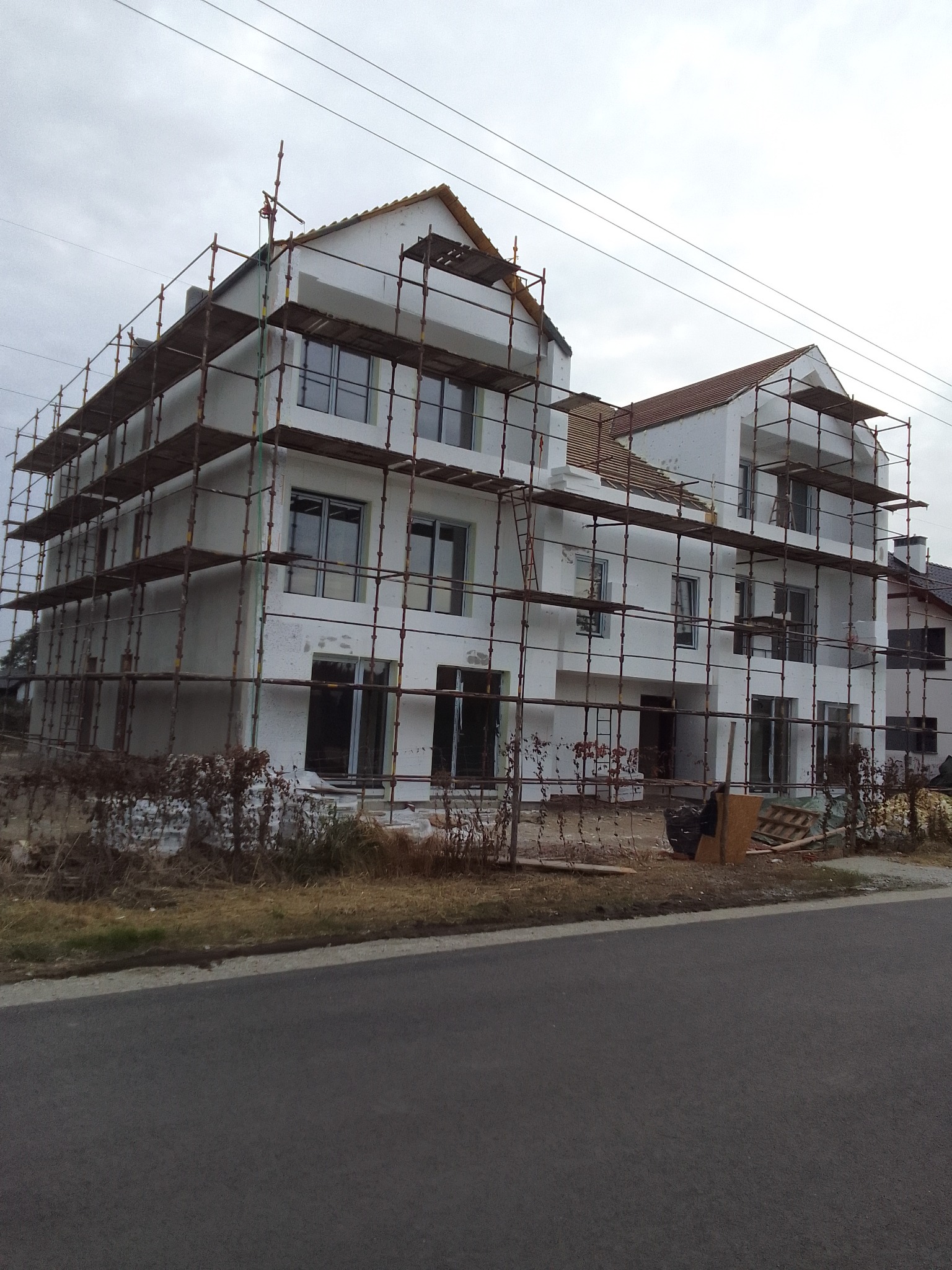 Budynek mieszkalny w trakcie prac wykończeniowych, z widocznymi rusztowaniami i świeżo nałożonym tynkiem, częściowo w kolorze białym, z oknami bez parapetów, w tle pochmurne niebo.