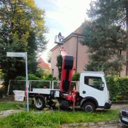 Drzewołazik Usługi Ogrodnicze Bartosz Nawojczyk - Ciężarówka z podnośnikiem koszowym na tle drzew i budynku, osoba w koszu na wysokości dachu, znak uliczny 'ul. Grabskiego'.