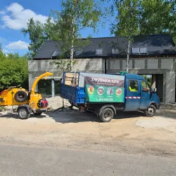 Żółty rębak do gałęzi i niebieska ciężarówka firmy Drzewołazik na tle lasu i budynku w trakcie budowy. Na ciężarówce reklama firmy.