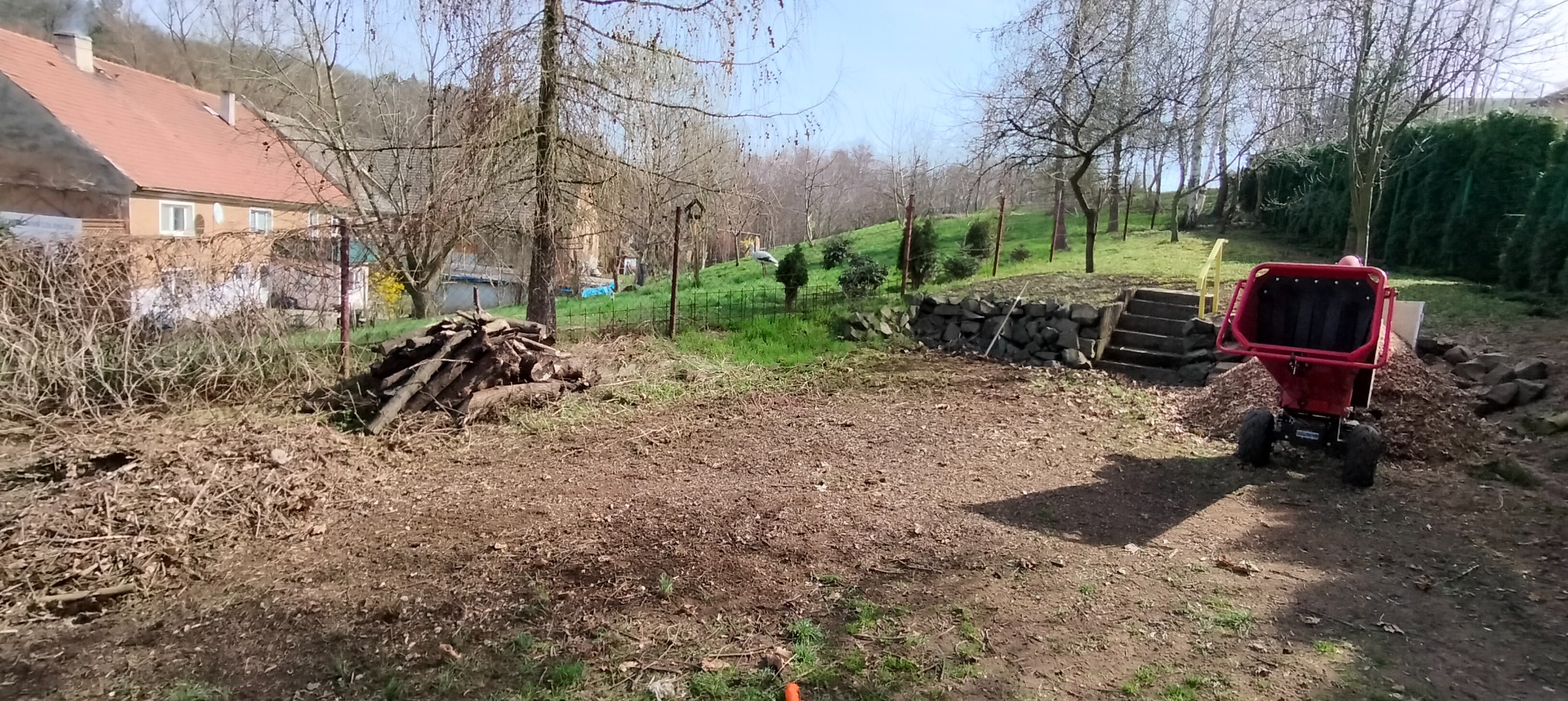 Teren po wycince drzew z ułożonymi gałęziami i rozdrabniarką do gałęzi, widoczne schody kamienne i trawa z zielenią w tle.