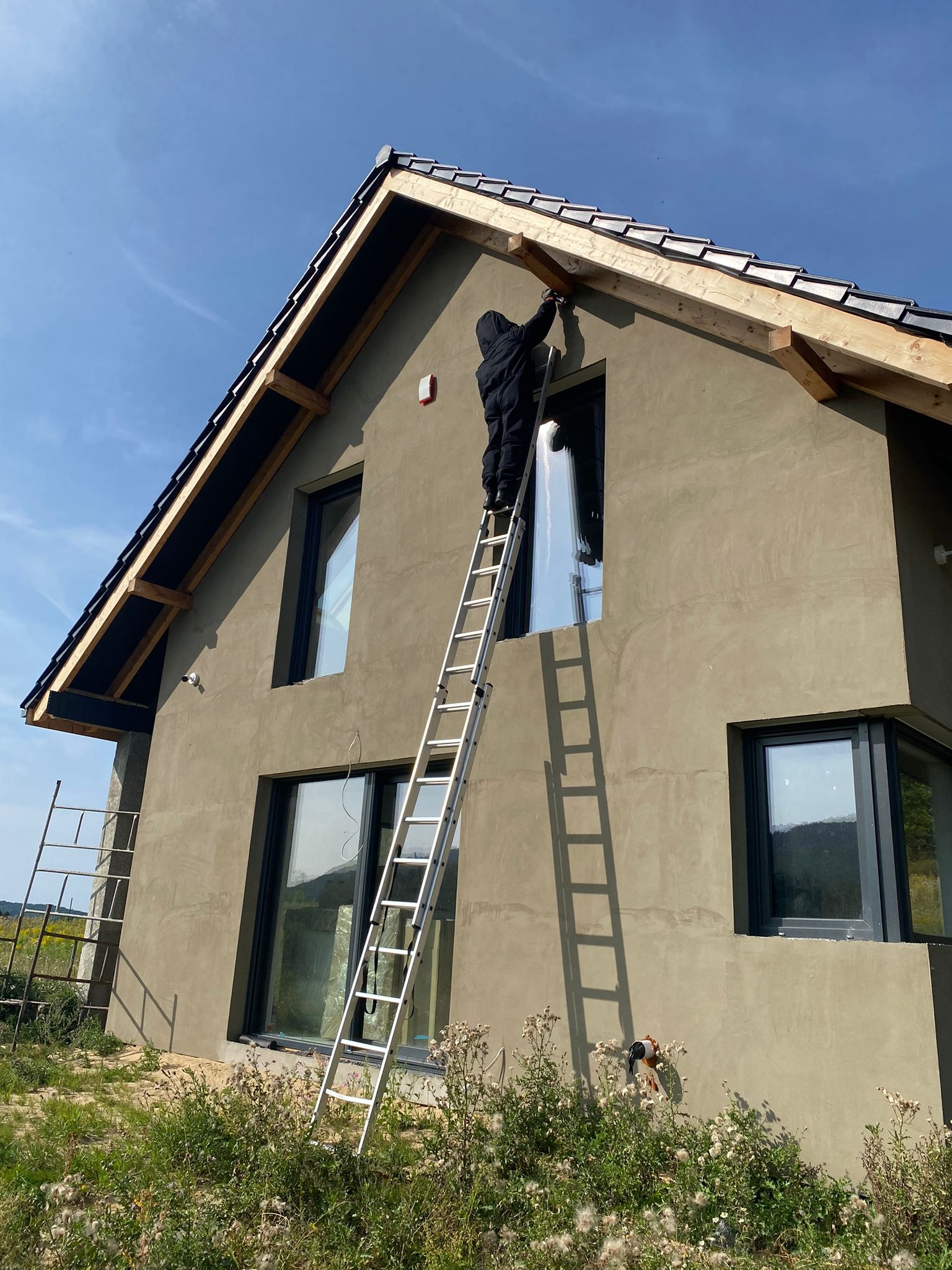 Pracownik w czarnym kombinezonie na aluminiowej drabinie, montujący elementy drewniane pod dachem nowo otynkowanego domu jednorodzinnego w słoneczny dzień.