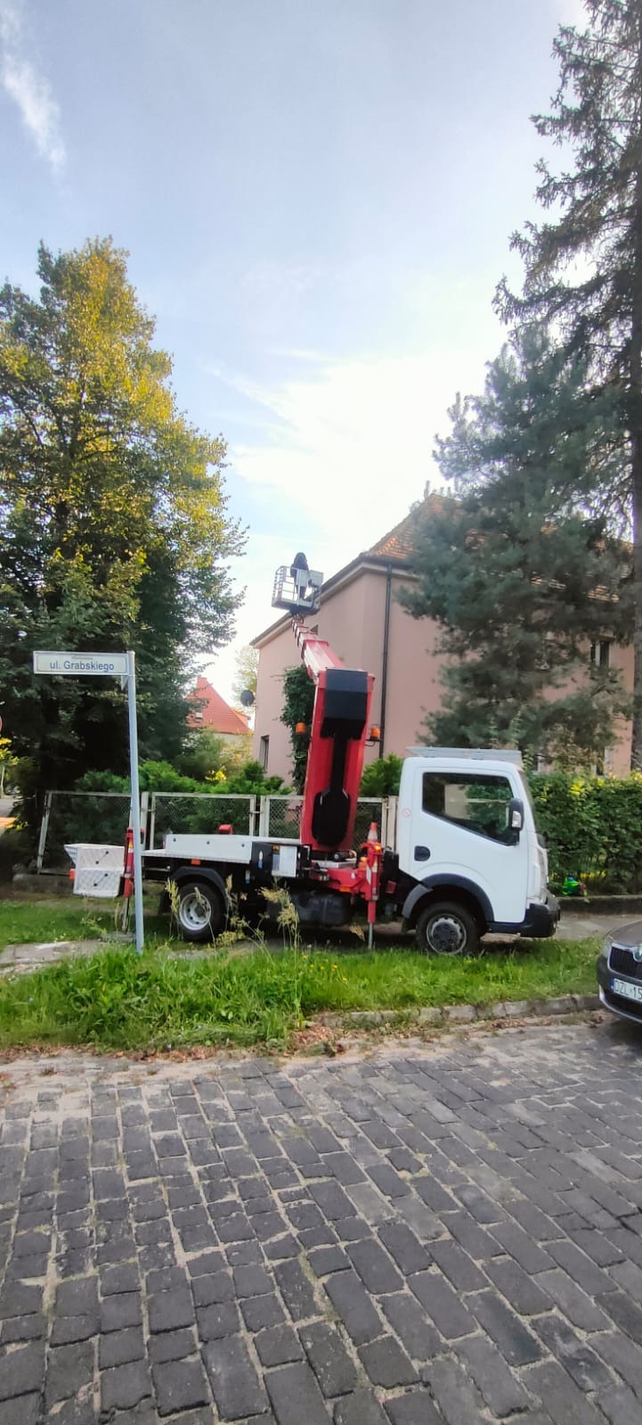 Ciężarówka z podnośnikiem koszowym na tle drzew i budynku, osoba w koszu na wysokości dachu, znak uliczny 'ul. Grabskiego'.