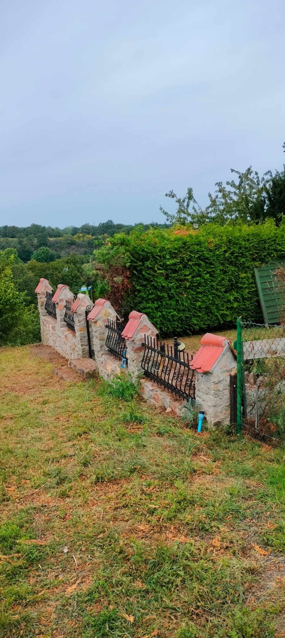 Kamienny murek z czerwonymi daszkami i metalowym ogrodzeniem, otoczony zielenią i drzewami w tle.
