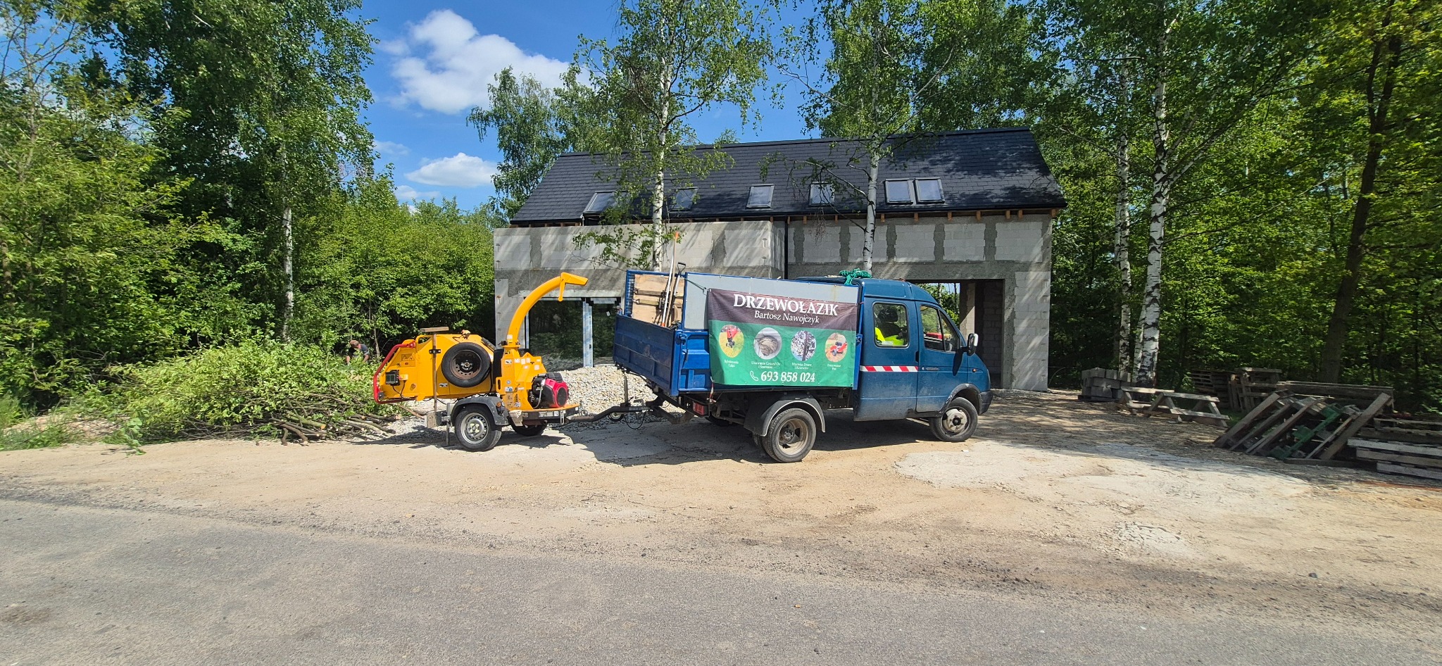 Żółty rębak do gałęzi i niebieska ciężarówka firmy Drzewołazik na tle lasu i budynku w trakcie budowy. Na ciężarówce reklama firmy.