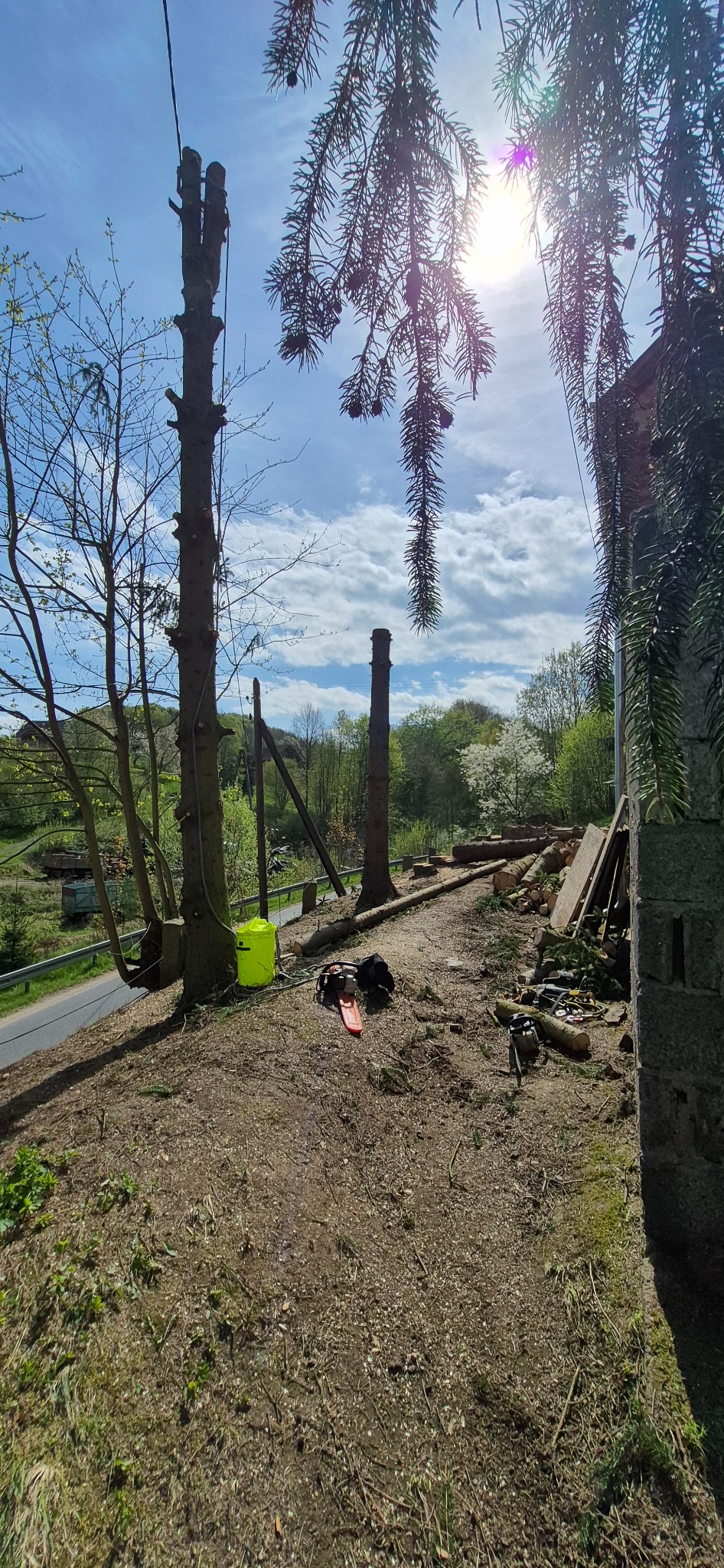 Teren po wycince drzew: pnie, ścięte gałęzie, piła łańcuchowa i sprzęt BHP. W tle błękitne niebo i zieleń drzew. Widok z góry.