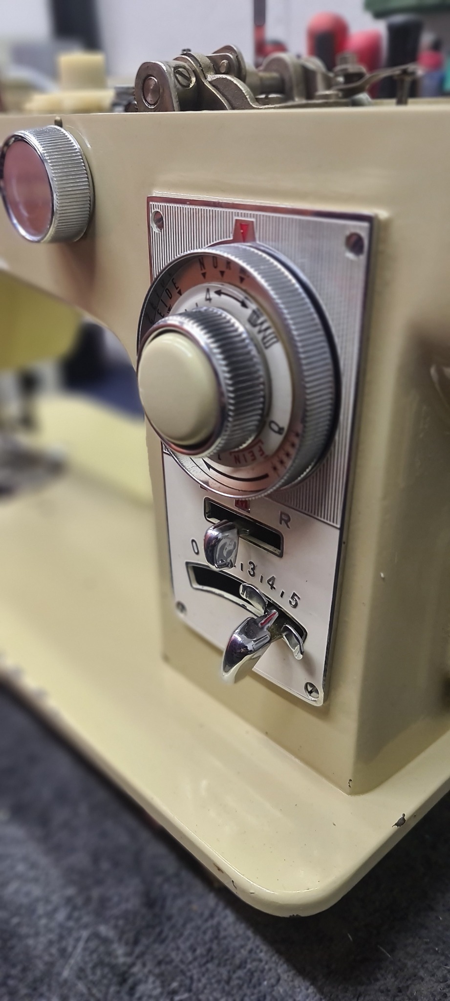 Zbliżenie na panel sterowania beżowej maszyny do szycia z chromowanymi pokrętłami i suwakami ustawionymi na wartościach. Widoczne oznaczenia 'NUR FEIDE' i skala od 0 do 5.