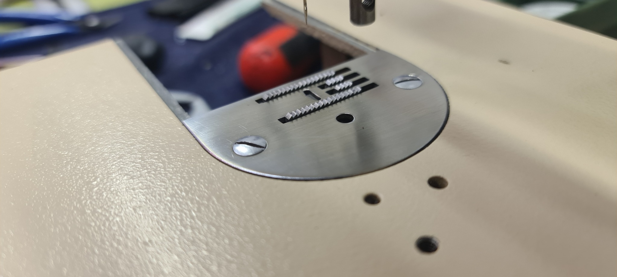 Zbliżenie na metalową płytkę ściegową maszyny do szycia z widoczną igłą i ząbkami transportera materiału.