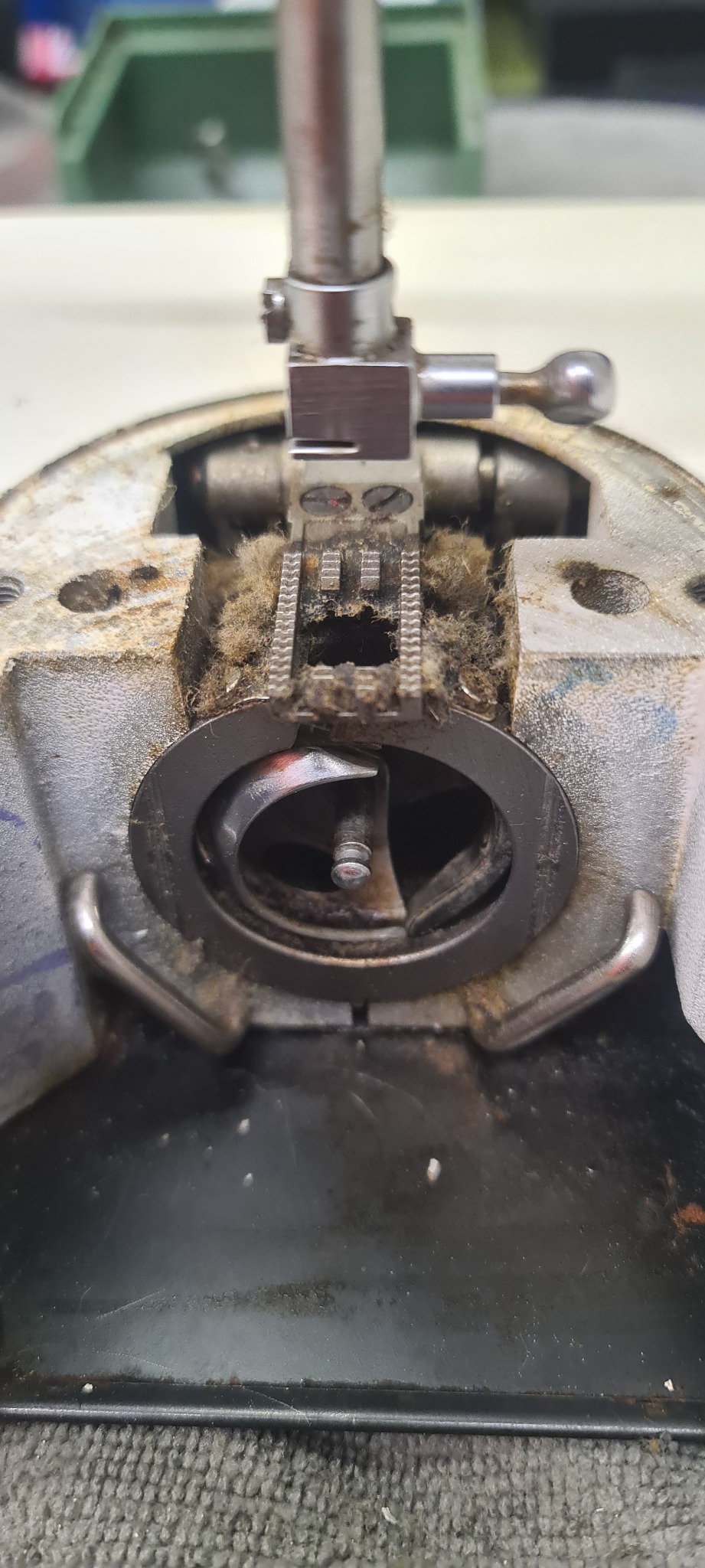 Detail wewnętrznych mechanizmów starej maszyny do szycia, widoczny z bliska obszar chwytacza i ząbków transportujących materiał, zanieczyszczony kurzem i resztkami nici.
