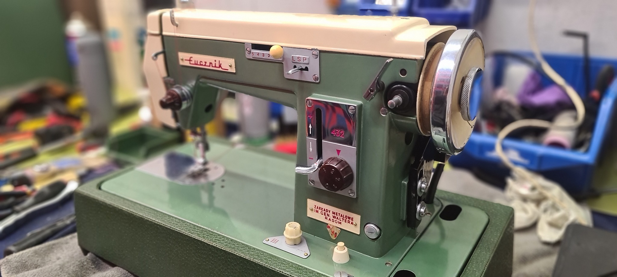 Antyczna maszyna do szycia Łucznik 432 w kolorze zieleni i beżu, z widocznym pokrętłem regulacji, napisem 'Łucznik' i tabliczką informacyjną Zakłady Metalowe Radom, na rozmytym tle.