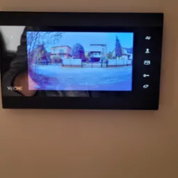 Czarny panel wideodomofonu Vrone zamontowany na beżowej ścianie, wyświetlający obraz z kamery zewnętrznej przedstawiający domy i ulicę w dzień.