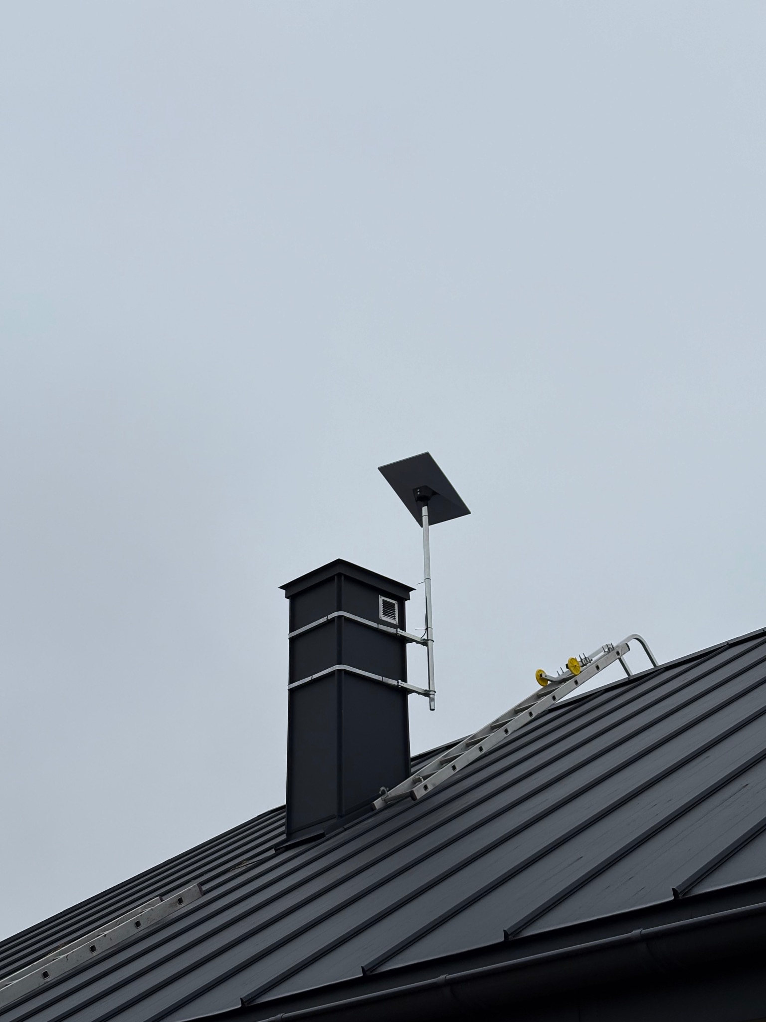 Antena Starlink zamontowana na dachu obok komina, drabina oparta o dach. Widok z dołu na szarym tle nieba.