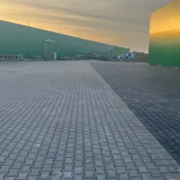 Rozległy plac wyłożony szarą kostką brukową typu holland, z widoczną linią podziału na jaśniejszą i ciemniejszą część, otoczony zielonymi halami magazynowymi w świetle zachodzącego słońca.