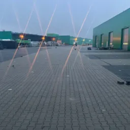 Rozległy plac wyłożony szarą kostką brukową w jodełkę, z widocznymi zielonymi budynkami magazynowymi w tle, paletami i zbiornikami, oraz czerwonym samochodem dostawczym z numerem telefonu po prawej...