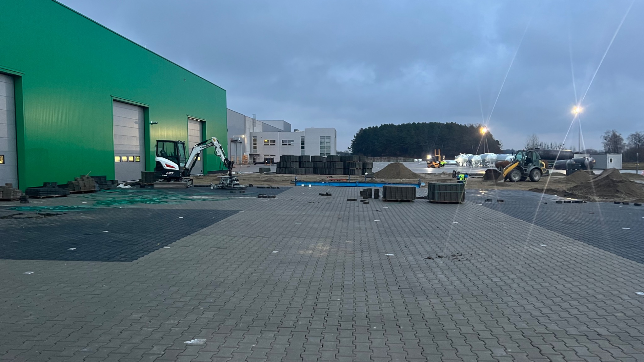 Układanie szarej kostki brukowej na dużej powierzchni placu przemysłowego, z widocznymi maszynami budowlanymi, stertami kostki i piasku, oraz budynkiem w tle, sfotografowane w pochmurny dzień.