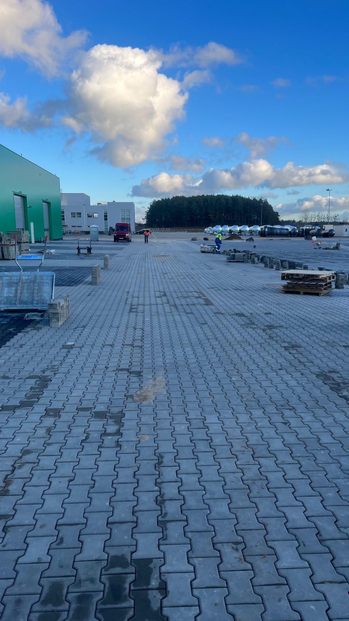 Układanie szarej kostki brukowej typu 'I' na dużej powierzchni placu przemysłowego, widoczni pracownicy w oddali, palety z kostką i wózek transportowy, błękitne niebo z chmurami w tle.