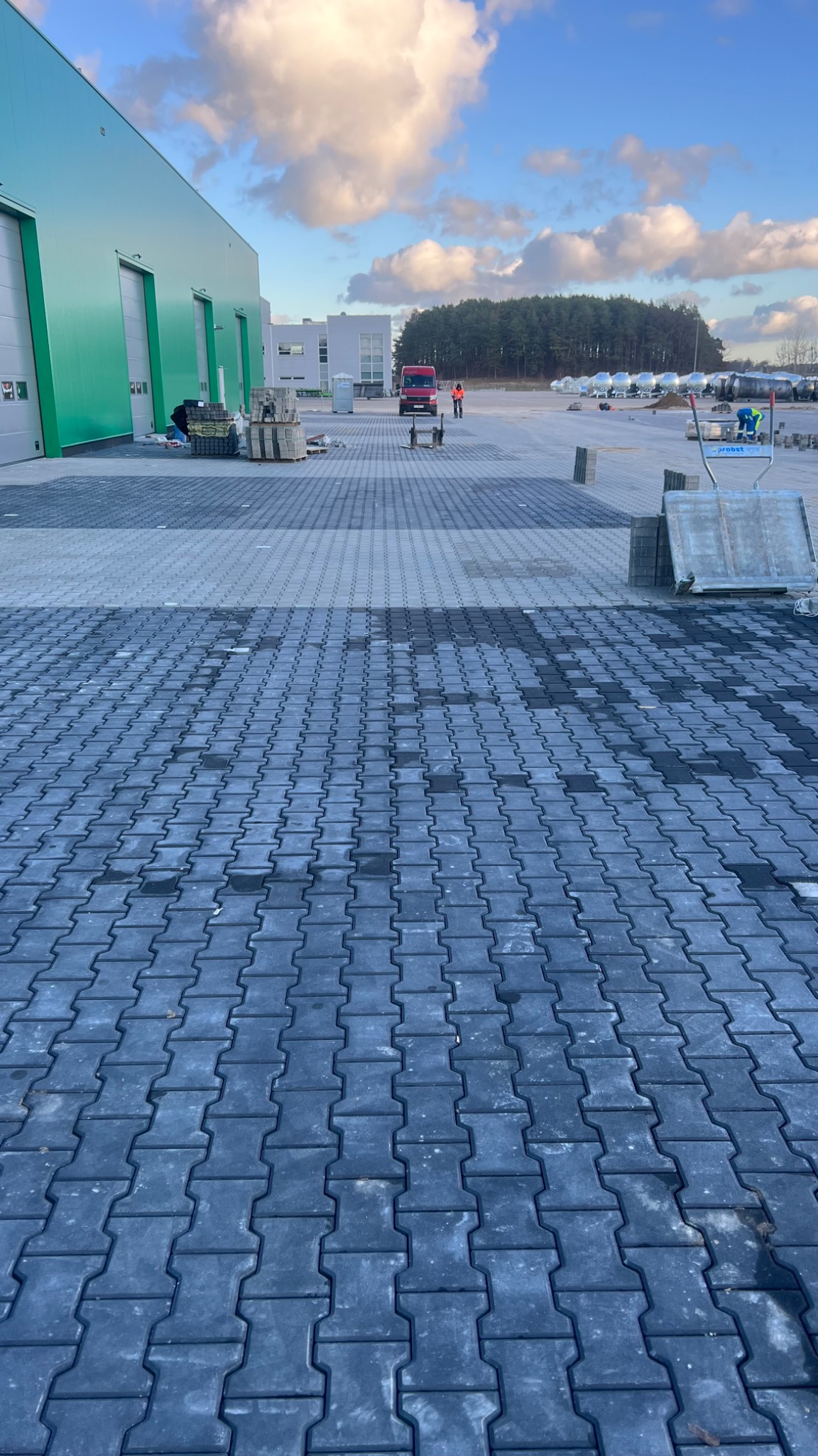 Częściowo ułożona, szara kostka brukowa na placu przemysłowym, z widocznymi pracownikami, paletami kostki i budynkiem magazynowym w tle pod błękitnym niebem.
